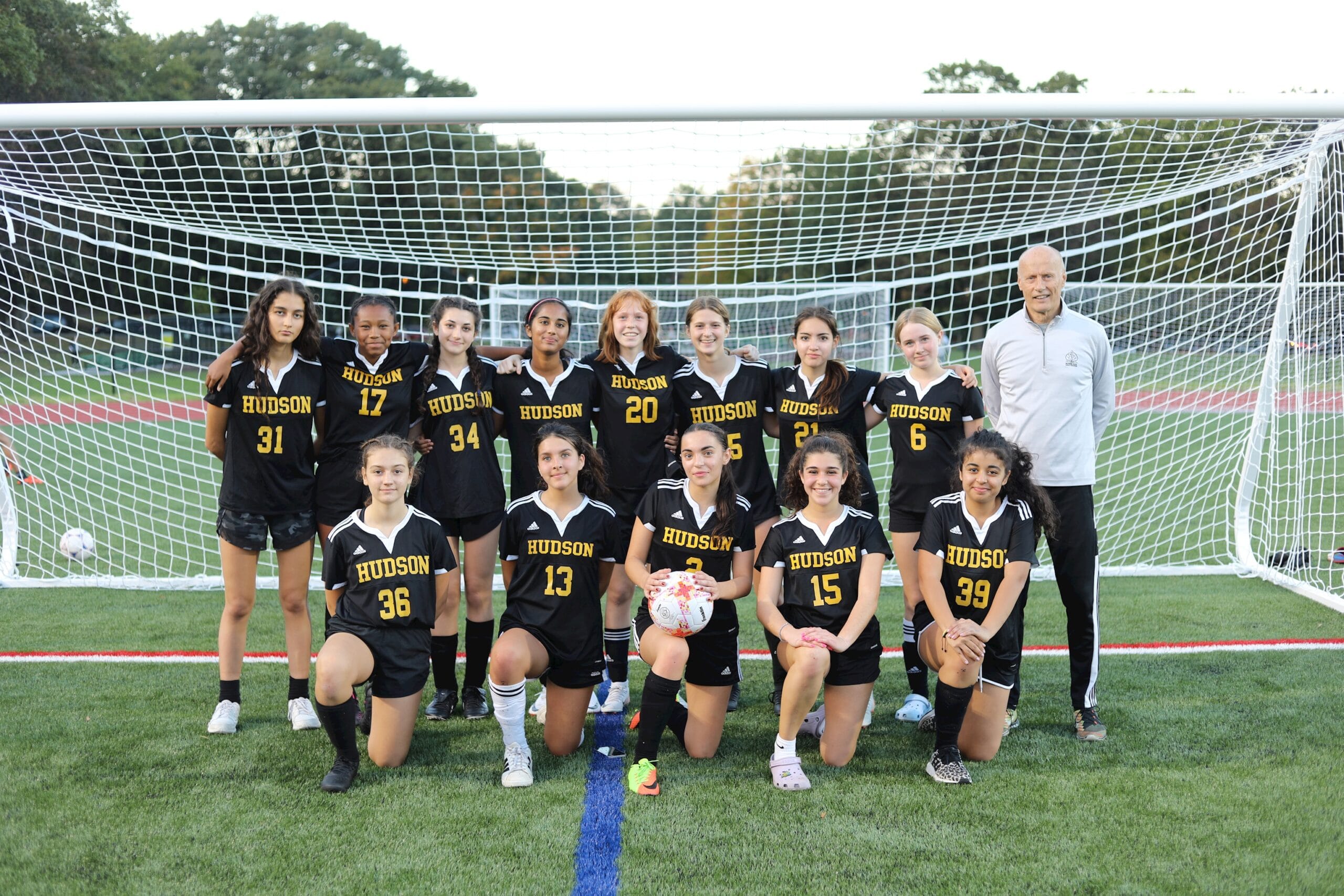 The Hudson School girls soccer team in black-and-gold uniforms posing with their coach in front of a goal