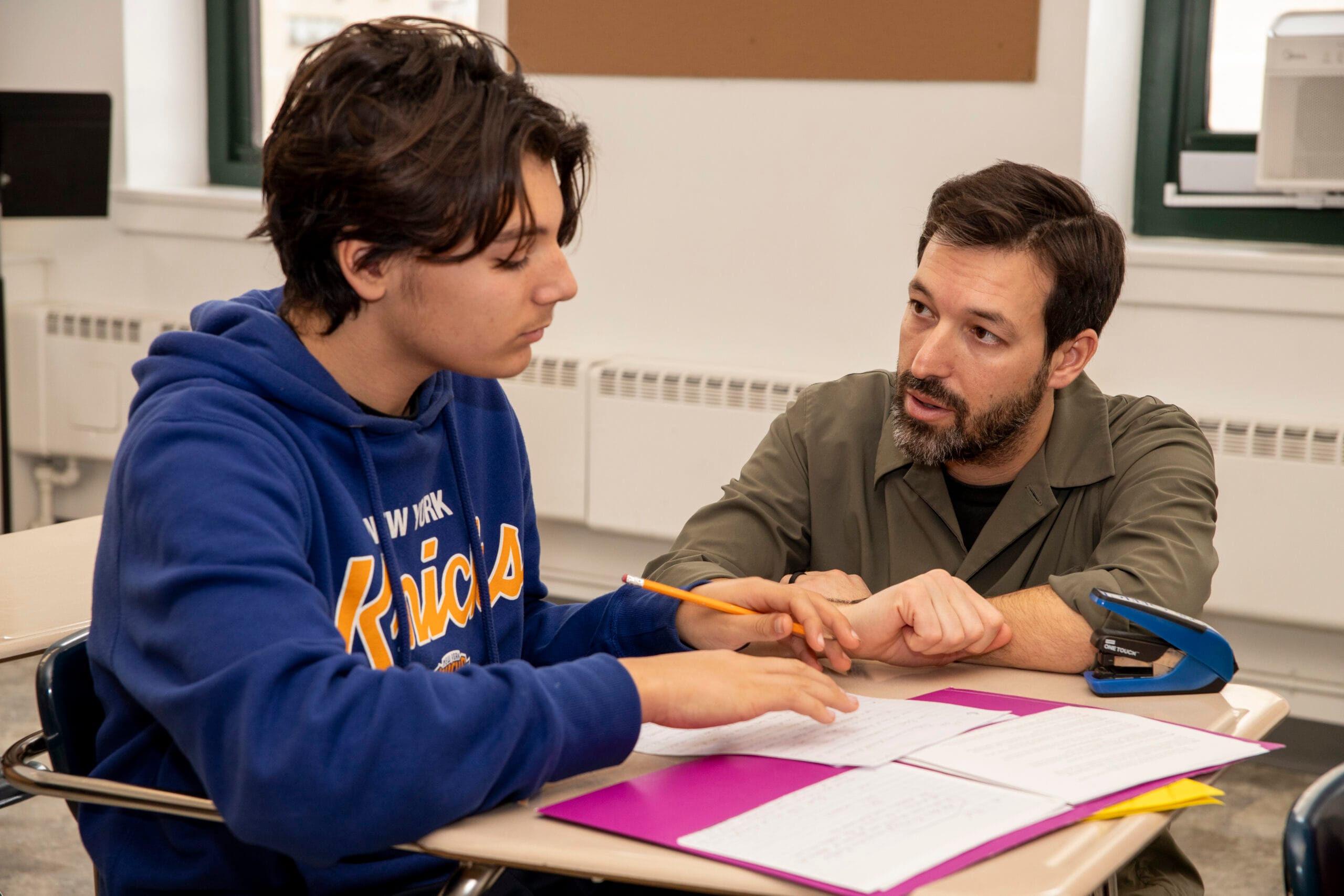 A Hudson School teacher working one-on-one with a student at a desk, reviewing written work together.