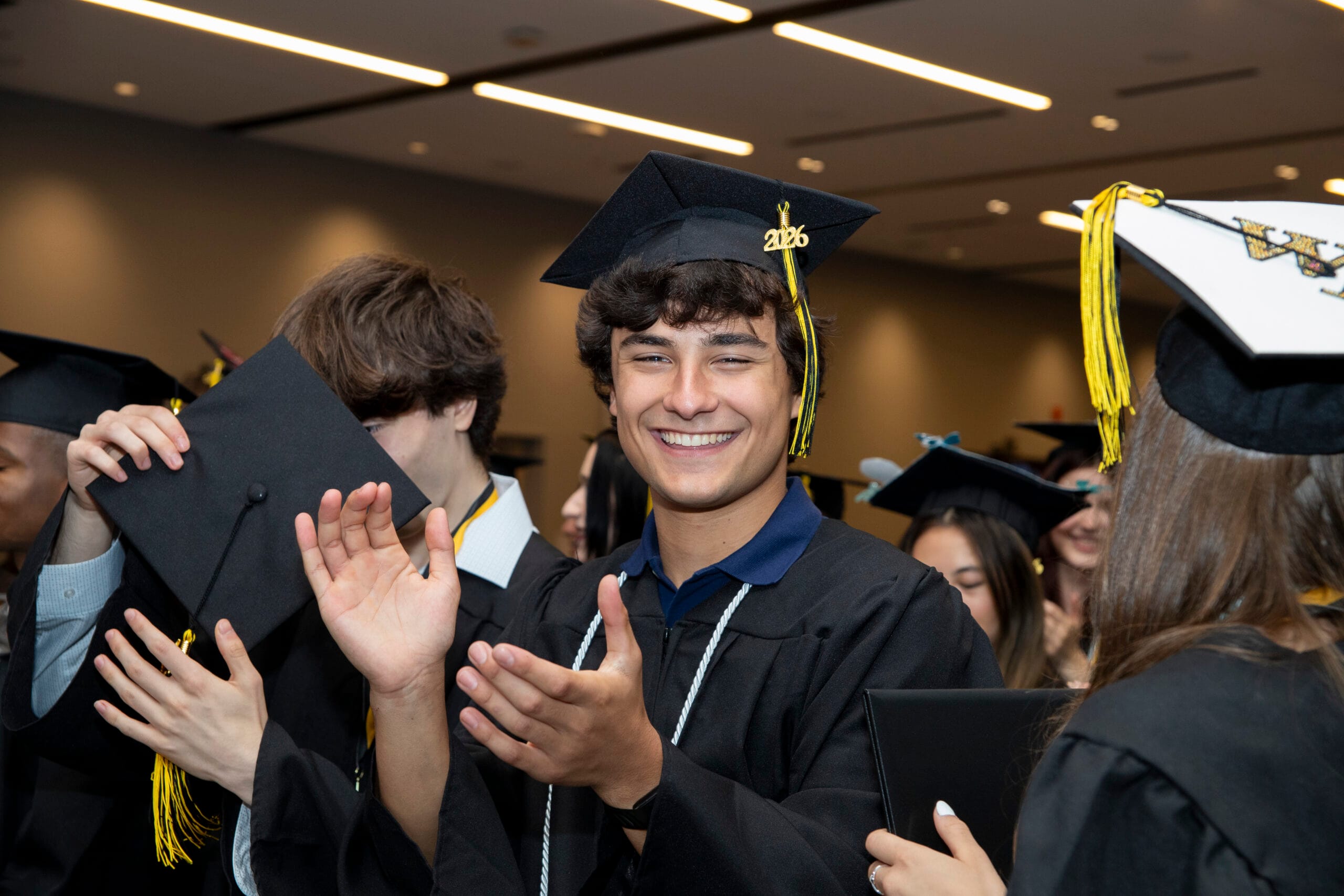 A smiling graduate with a 2026 tassel applauds classmates at commencement