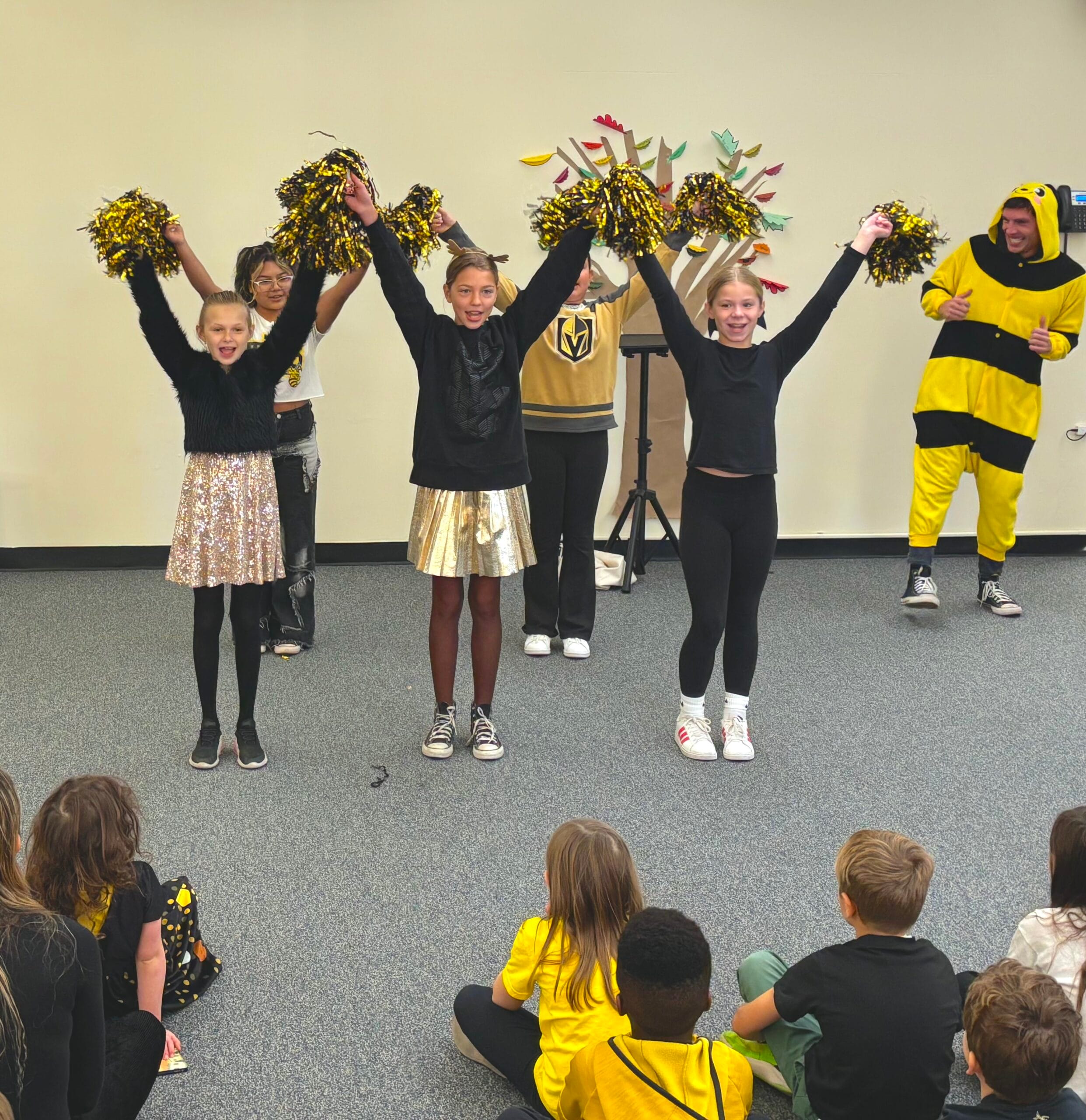 Students in black and gold shake pompoms during a Hudson School spirit performance as younger students watch